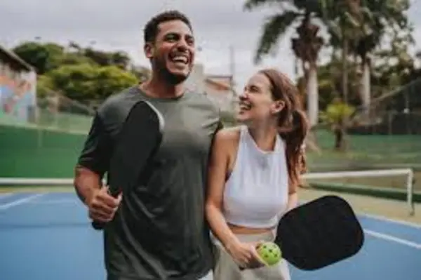 A man and woman stand together on a blue tennis court, each holding a paddle, smiling in a casual, outdoor setting.