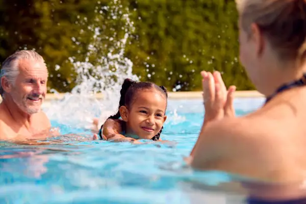 A child is swimming in a pool, splashing water, while an adult engages and encourages from the side. Bright, sunny day, lush greenery in background.